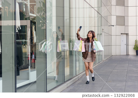 Asian woman enjoying shopping, happy woman holding colorful fashion bags near mall, celebrating lifestyle, people enjoying black friday sales, wearing stylish outfit, taking selfie with phone. 132922008