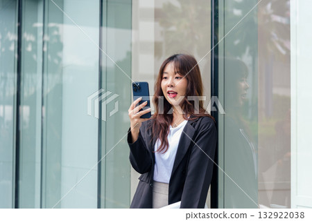 Young Asian business woman in suit on street, busy with phone communication for work, leaning against modern glass building exterior. 132922038