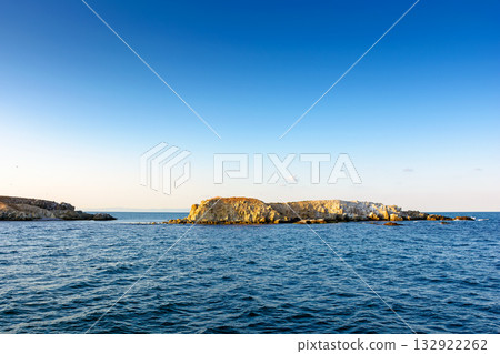 rock island view on black sea in evening light. rough landscape with calm waves for summer adventures background at the sea under blue sky with clouds rock island view on black sea in evening light. rough landscape with calm waves for summer adventures background at the sea under blue sky with clouds 132922262