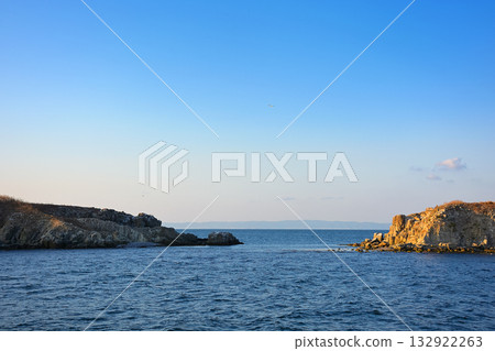 rock island view on black sea in evening light. rough landscape with calm waves for summer adventures background at the sea under blue sky with clouds rock island view on black sea in evening light. rough landscape with calm waves for summer adventures background at the sea under blue sky with clouds 132922263