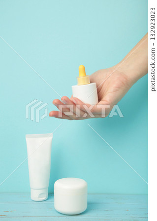 Set of various cosmetic products in blank packaging, creams in jars, serum in bottle on blue background. Woman holding bottle of serum Set of various cosmetic products in blank packaging, creams in jars, serum in bottle on blue background. Woman holding bottle of serum 132923023