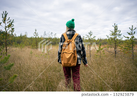 Rearview of woman with backpack walk through marshy terrain among tall grass under overcast sky Rearview of woman with backpack walk through marshy terrain among tall grass under overcast sky 132923879
