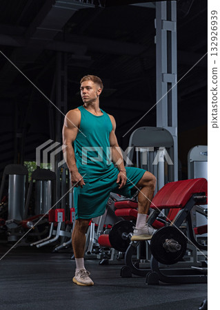 fit man stands in a gym one leg resting on gym equipment showcasing toned muscles. He appears focused and ready for his workout surrounded by exercise machines. fit man stands in a gym one leg resting on gym equipment showcasing toned muscles. He appears focused and ready for his workout surrounded by exercise machines. 132926939