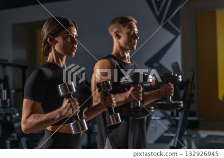 man and a woman are in a gym each holding dumbbells and performing hammer curls. They focus on their workout training together in the evening for strength and fitness. 132926945