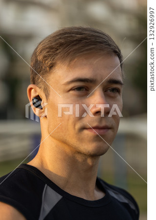 male athlete in a black sleeveless shirt listens to music with headphones while getting ready for an evening workout at a sports ground. The sun sets in the background. male athlete in a black sleeveless shirt listens to music with headphones while getting ready for an evening workout at a sports ground. The sun sets in the background. 132926997