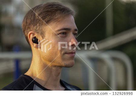 focused male athlete wearing headphones stands on a sports ground getting ready for training. He appears engaged in his workout routine as he enjoys music. 132926998