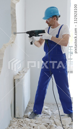 Man construction worker demolishing concrete wall with rotary hammer, wearing protective safety gear, generating dust and debris during home renovation work 132928100