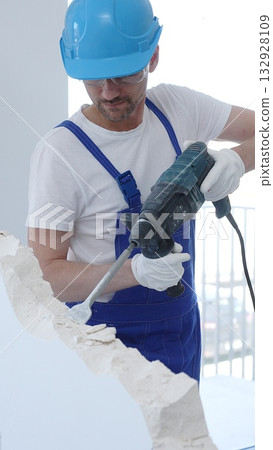 Male construction worker demolishing concrete wall with rotary hammer, wearing protective safety overalls is generating dust and debris during renovation project. Vertical portrait view Male construction worker demolishing concrete wall with rotary hammer, wearing protective safety overalls is generating dust and debris during renovation project. Vertical portrait view 132928109