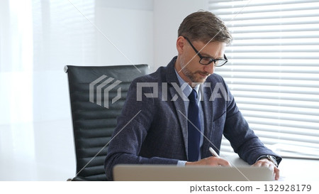 Senior businessman wearing suit, tail and black glasses is concentrating while writing on a document at his desk in a bright, modern office, demonstrating professionalism. Business people concept Senior businessman wearing suit, tail and black glasses is concentrating while writing on a document at his desk in a bright, modern office, demonstrating professionalism. Business people concept 132928179