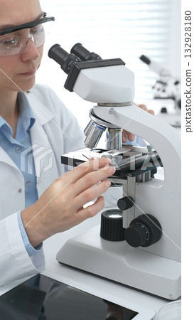 Female scientist wearing protective gear analyzing test samples under microscope in contemporary research environment. Medicine, healthcare and science concept 132928180