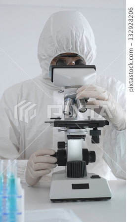 A medical virologist in protective gear examines viral samples under a microscope while conducting research in a laboratory setting 132928206