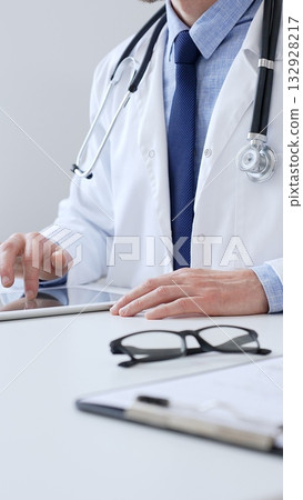 Adult male doctor wearing lab coat and stethoscope using digital tablet while sitting at desk in medical office, with medical chart and pen in the foreground, close up. Medicine Adult male doctor wearing lab coat and stethoscope using digital tablet while sitting at desk in medical office, with medical chart and pen in the foreground, close up. Medicine 132928217