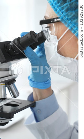 Female scientist in blue gloves and mask using microscope in laboratory setting, conducting important research and analysis, close-up. Medicine and science concept 132928230