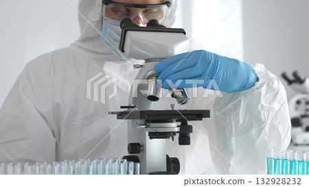 Medical virologist in protective hazmat gear examining viral samples under a microscope while conducting research in a sterile laboratory environment Medical virologist in protective hazmat gear examining viral samples under a microscope while conducting research in a sterile laboratory environment 132928232