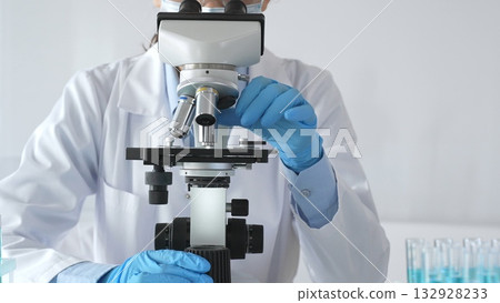 A female scientist in protective gear adjusts a microscope lens and accurately analyzes medical research samples in a sterile laboratory setting 132928233