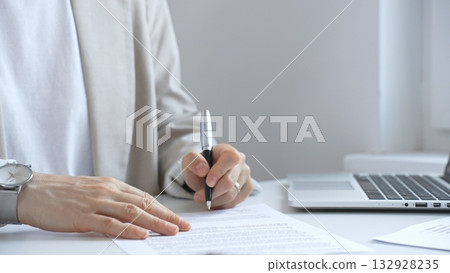 Businesswoman signing a contract at office desk, surrounded by a laptop and documents, exemplifies focus and commitment. Business people Businesswoman signing a contract at office desk, surrounded by a laptop and documents, exemplifies focus and commitment. Business people 132928235