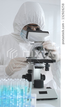 Medical scientist wearing protective suit using microscope while analyzing samples, conducting research in laboratory environment. Close up of test tubes with blue liquid 132928258