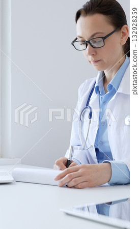 Adult doctor woman completing healthcare paperwork, clipboard positioned near laptop and stethoscope in well lit clinical workspace by window. Medicine and health care 132928259