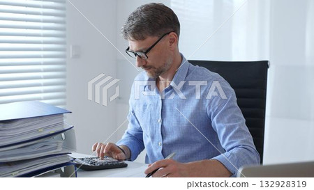 Adult businessman wearing blue sleek shirt and glasses, is thinking and concentrating while working at the desk in a bright, modern office, demonstrating professionalism and efficiency. Business Adult businessman wearing blue sleek shirt and glasses, is thinking and concentrating while working at the desk in a bright, modern office, demonstrating professionalism and efficiency. Business 132928319