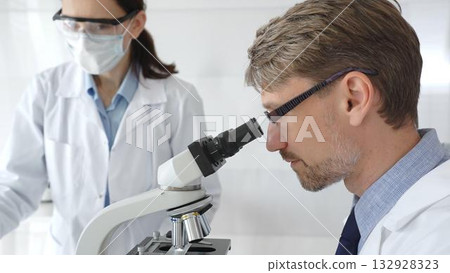 A researcher wearing glasses examines a sample under a microscope, a lab technician wearing a protective mask stands nearby, both in white lab coats during the study. Medicine and Science A researcher wearing glasses examines a sample under a microscope, a lab technician wearing a protective mask stands nearby, both in white lab coats during the study. Medicine and Science 132928323