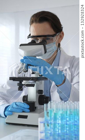 Female scientist examining microscope slide, wearing protective gear in laboratory, researching pharmaceutical breakthrough with blue tinted test tube specimens Female scientist examining microscope slide, wearing protective gear in laboratory, researching pharmaceutical breakthrough with blue tinted test tube specimens 132928324