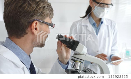 Scientific professional examining specimen through microscope, laboratory assistant nearby wearing protective mask, both dressed in white lab coats during research process. Medicine and science 132928325