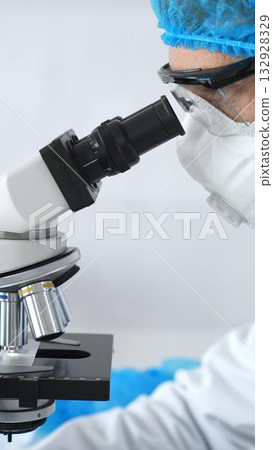 Medical scientist senior man wearing protective mask, glasses and bouffant cap using microscope doing scientific research in a laboratory. Medicine and science concept Medical scientist senior man wearing protective mask, glasses and bouffant cap using microscope doing scientific research in a laboratory. Medicine and science concept 132928329