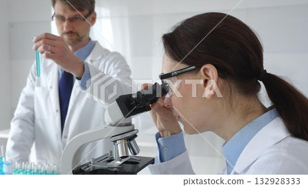A female scientist wearing protective glasses looks into a microscope and conducts a medical research in a sterile laboratory while her male colleague works in the background. Medicine and science 132928333