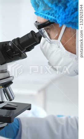 Medical scientist senior man wearing protective mask, glasses and bouffant cap using microscope doing scientific research in a laboratory. Medicine and science concept Medical scientist senior man wearing protective mask, glasses and bouffant cap using microscope doing scientific research in a laboratory. Medicine and science concept 132928344