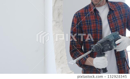 Male construction worker wearing red checkered shirt and protective white gloves is demolishing white wall with rotary hammer drill and generating dust during renovation project, closeup view Male construction worker wearing red checkered shirt and protective white gloves is demolishing white wall with rotary hammer drill and generating dust during renovation project, closeup view 132928345
