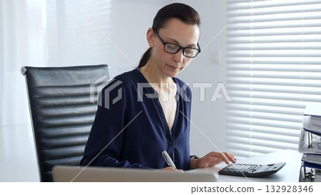 Professional woman accountant wearing blue blouse and black glasses, is working at office desk, calculating finances, writing notes, managing business documents. Audit and taxes in business Professional woman accountant wearing blue blouse and black glasses, is working at office desk, calculating finances, writing notes, managing business documents. Audit and taxes in business 132928346