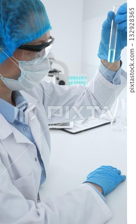 Medical science researcher wearing blue protective gear of cap, gloves and googles, is examining liquid in test tube in laboratory workspace. Medicine and health care concept Medical science researcher wearing blue protective gear of cap, gloves and googles, is examining liquid in test tube in laboratory workspace. Medicine and health care concept 132928365