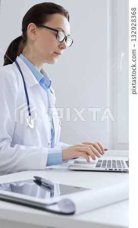 Female doctor is typing on a laptop in a bright medical office, showcasing healthcare technology and medical documentation. Medicine and health care concept Female doctor is typing on a laptop in a bright medical office, showcasing healthcare technology and medical documentation. Medicine and health care concept 132928368
