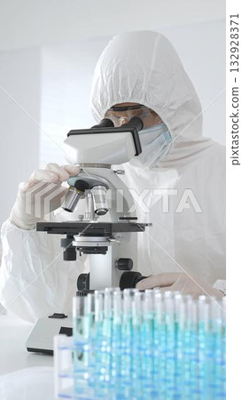 A medical virologist in protective gear examines viral samples under a microscope while conducting research in a laboratory setting 132928371