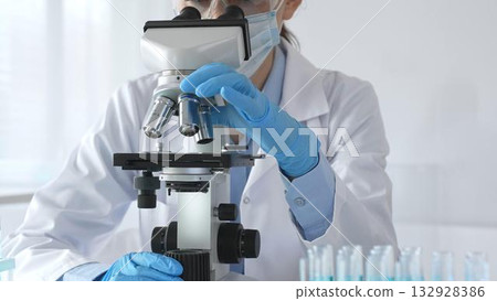 A female scientist in protective gear adjusts a microscope lens and accurately analyzes medical research samples in a sterile laboratory setting A female scientist in protective gear adjusts a microscope lens and accurately analyzes medical research samples in a sterile laboratory setting 132928386