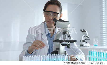 Female scientist in lab coat and safety glasses using microscope, analyzing samples in test tubes in laboratory, conducting scientific research Female scientist in lab coat and safety glasses using microscope, analyzing samples in test tubes in laboratory, conducting scientific research 132928399