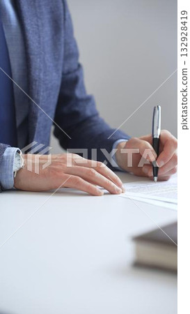Close up of businessman's hands signing a contract with elegant pen, wearing a blue suit and a wristwatch, sitting at a white desk. Business people concept 132928419