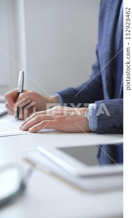 Closeup of businessman hands signing a contract with elegant pen, wearing a blue suit and a wristwatch, sitting at a white desk in office. Business people concept 132928462