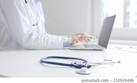 Closeup of blue stethoscope is lying on the table near female doctor working on laptop, highlighting digital tools transforming healthcare diagnostics and patient documentation. Medicine concept 132928486
