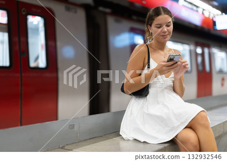 Positive young girl sitting on subway platform holding phone in hand 132932546