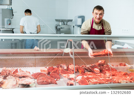 Butcher in an apron offers to buy fresh meat in grocery store section 132932680