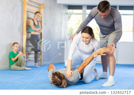 Woman is learning how to grab opponent's hand in prone position during self-defense class, in background girl and boy watch sparring Woman is learning how to grab opponent's hand in prone position during self-defense class, in background girl and boy watch sparring 132932717