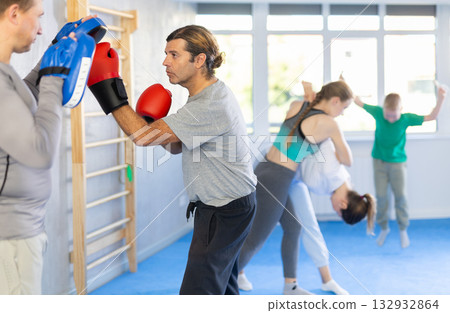Man in boxing gloves practices punches with man in mitts in sport club during group training Man in boxing gloves practices punches with man in mitts in sport club during group training 132932864