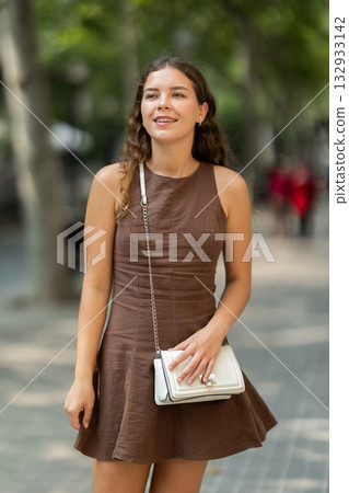 Beautiful stylish young girl in dress with white handbag walking and posing on city street on summer day 132933142