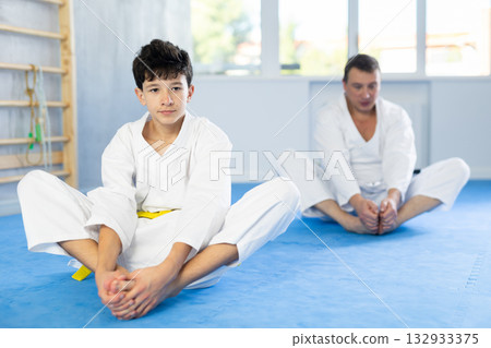 Teenager boy and coach in kimono sits in butterfly pose and practices stretching in sport gym 132933375