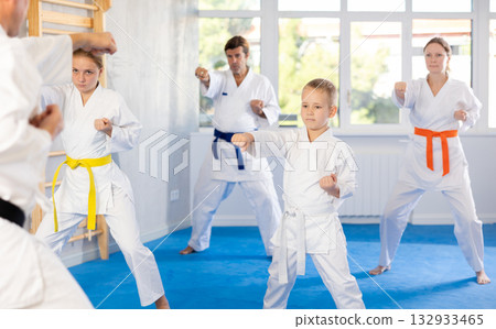 Boy with his family in kimonos and colored belts practicing karate with punches during group martial arts class in gym, accompanied by trainer Boy with his family in kimonos and colored belts practicing karate with punches during group martial arts class in gym, accompanied by trainer 132933465