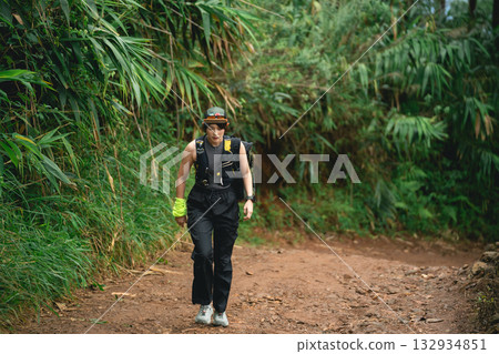 Young adult hiking on a scenic dirt trail surrounded by lush greenery and tropical plants, wearing hiking gear and enjoying nature in a serene environment 132934851