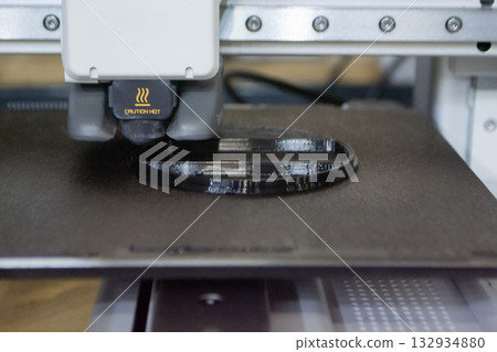 3D printer close up. Working 3D printer in process of printing object from molten black plastic. 3D printer creating model by flowing liquid plastic from extruder of printer. 3D printing technology 132934880