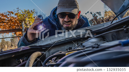 Man inspecting car engine with flashlight during early evening Man inspecting car engine with flashlight during early evening 132935035