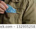 Hand putting Canadian money in pocket.person placing a blue Canadian five-dollar banknote into the chest pocket of a khaki jacket, symbolizing savings, cash handling, or financial security. 132935258